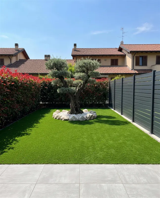 Giardino di 50 m² in prato sintetico a Nerviano con ulivo al centro dove prende sole tutto il giorno, sassi bianchi intorno al tronco