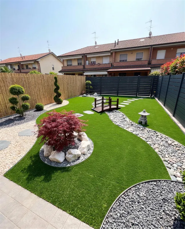 Giardino di 100 m² in prato sintetico a Varese con fiume di ciottoli che divide il giardino senza chiuderlo, ponte e lastre