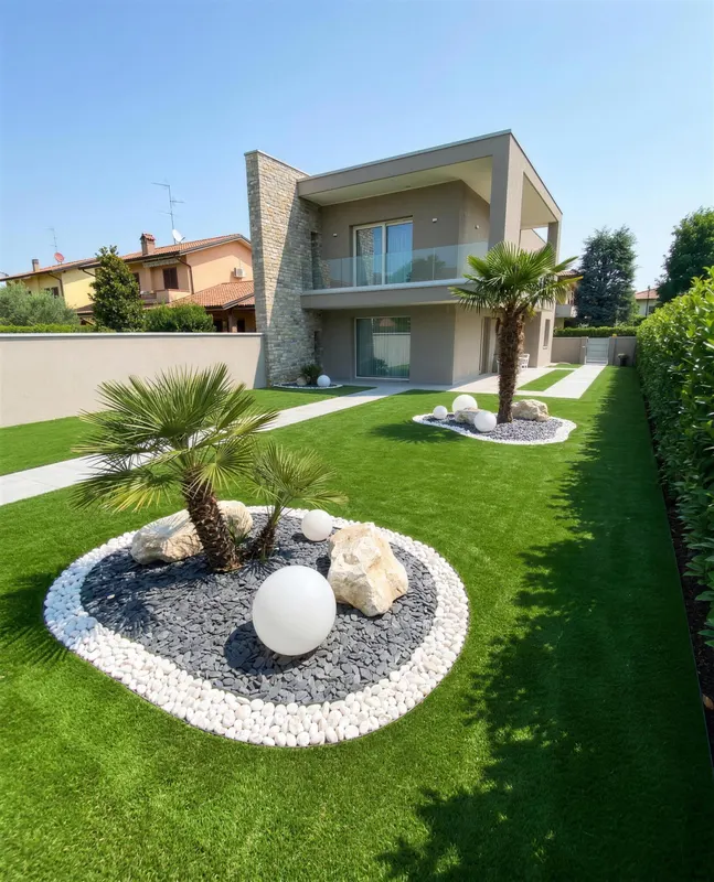 Giardino di 200 m² in prato sintetico a Brescia con aiuole a isola col bordo di ciottoli bianchi, palme dentro le aiuole così il prato resta libero